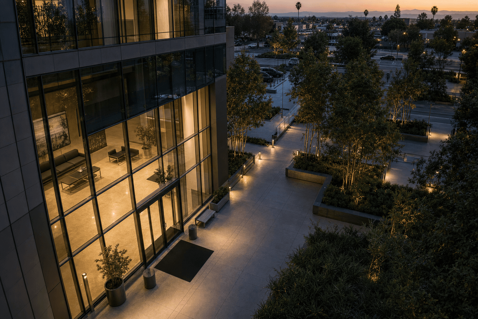 High neutral viewpoint over an empty, warmly lit office lobby and landscaped walkway at dusk